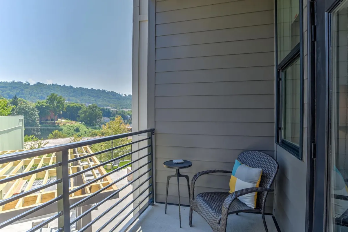 An inviting balcony scene with a wicker chair and a small side table set against a balcony railing. The view extends to a distant landscape, suggesting city views with hints of surrounding natural features. The setup is perfect for sipping coffee, reading, or quietly enjoying the outdoors.