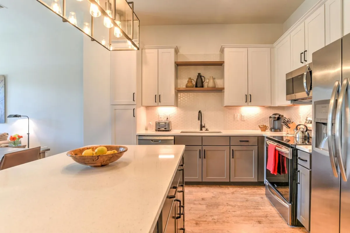 A modern kitchen with a central island topped with a bowl of fresh fruit, sleek appliances, and open shelving adorned with decorative objects. The space is designed for both functionality and entertaining, with ample room for cooking and social gatherings.
