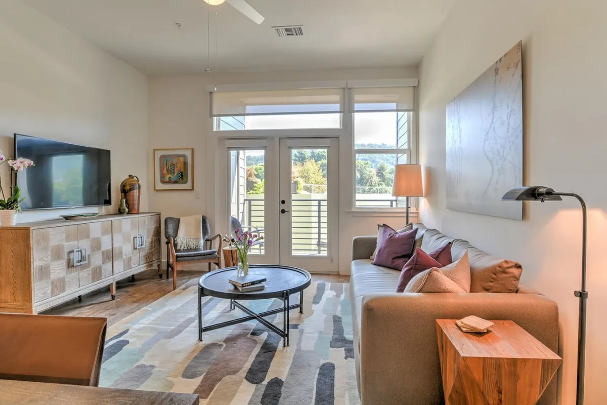 A modern, comfortable living room with a sofa, coffee table, television, striking wall art, and a flower-filled vase. The space hints at access to a balcony where couples can enjoy sunset views, tying in the section’s mention of private balcony moments.