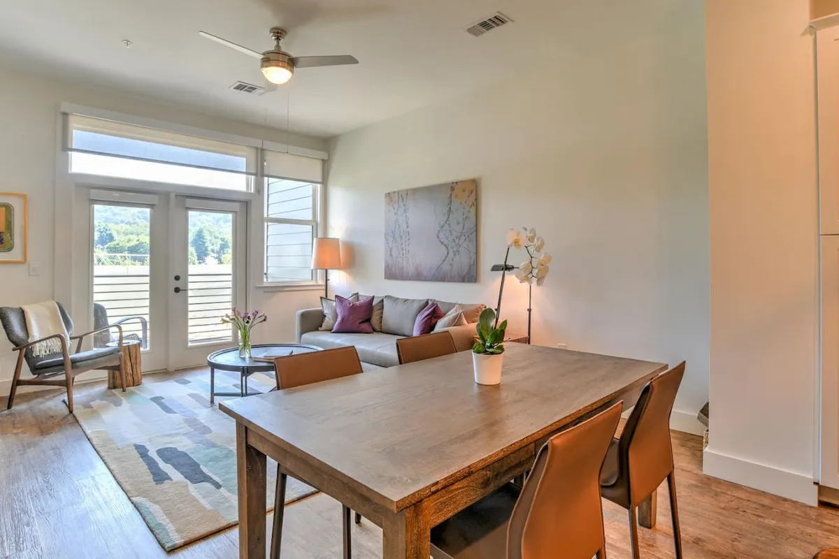 A cozy scene showing a dining table adjacent to a plush sofa with colorful cushions, all bathed in natural light streaming through doors that open to an outdoor view. The setup highlights both dining and lounging areas within one open, unified space.