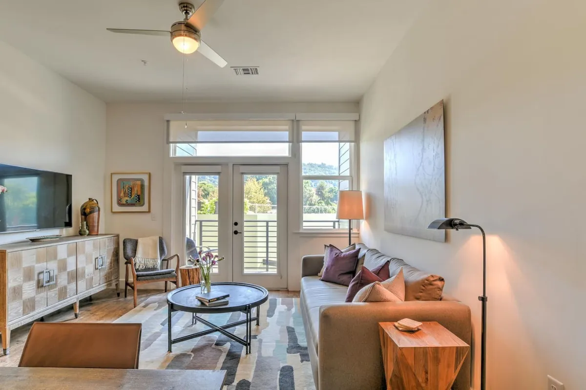 A stylish, well-lit living room featuring a comfortable sofa, coffee table, television, modern artwork on the wall, and a view out to the balcony. Natural light pours in through large glass doors, illuminating the fresh, contemporary furnishings and creating a welcoming atmosphere.