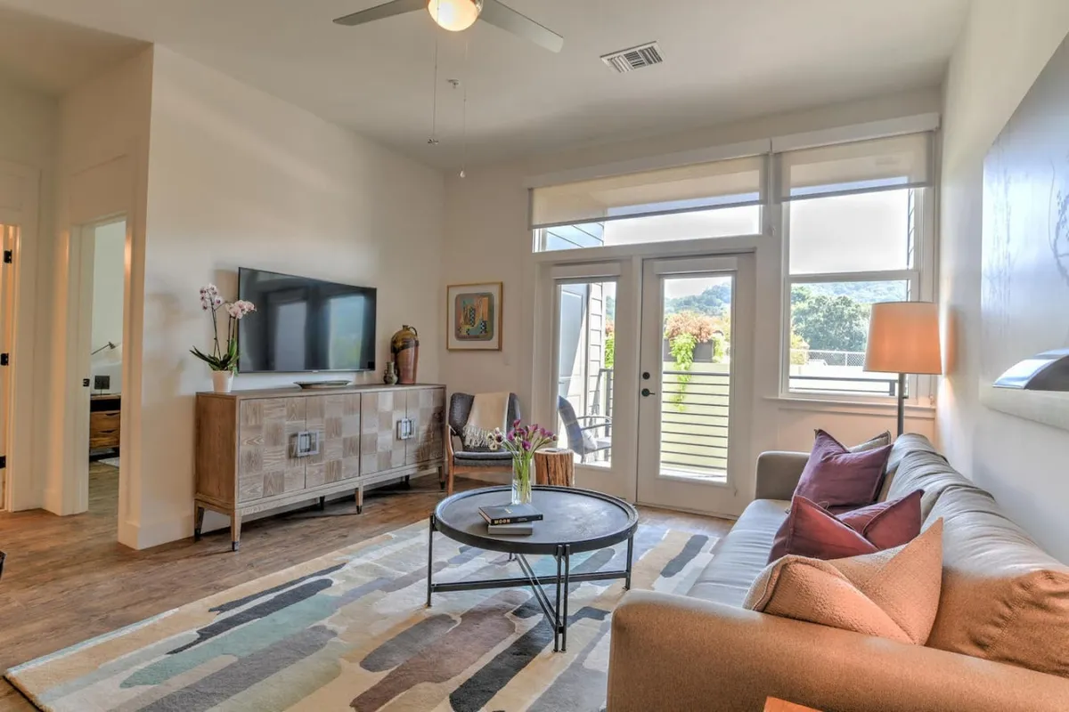 An inviting, sunlit open-concept living area featuring a plush sofa, sleek coffee table, smart TV, and vibrant decorative plants, all framed by expansive windows that fill the space with natural light. The modern decor and cozy atmosphere set the perfect stage for relaxing mornings or lively evenings with friends and family.