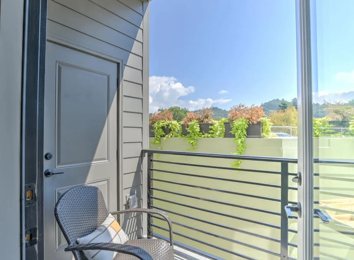 A tranquil balcony with a comfortable chair, a door leading inside, and lush planters adding greenery. The scene is bathed in soft natural light, suggesting a peaceful spot for morning coffee or quiet reflection, and subtly integrates the indoor-outdoor connection.
