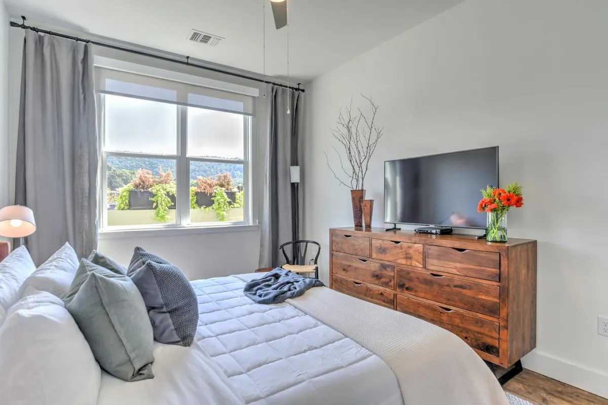 A tranquil bedroom featuring a bed with decorative pillows, a wooden dresser, television, and a vase with fresh flowers. A window provides a view outside, filling the space with natural light. The setting emphasizes comfort and peacefulness.